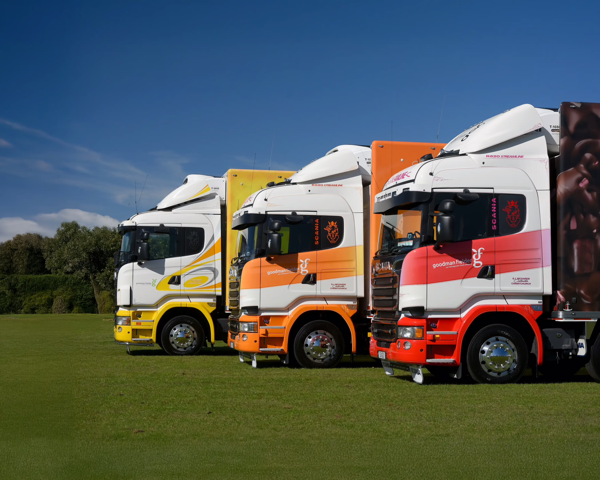 Custom vehicle wraps by Timaru Signs on Goodman Fielder Scania trucks in orange, yellow, and red color schemes on a green field.