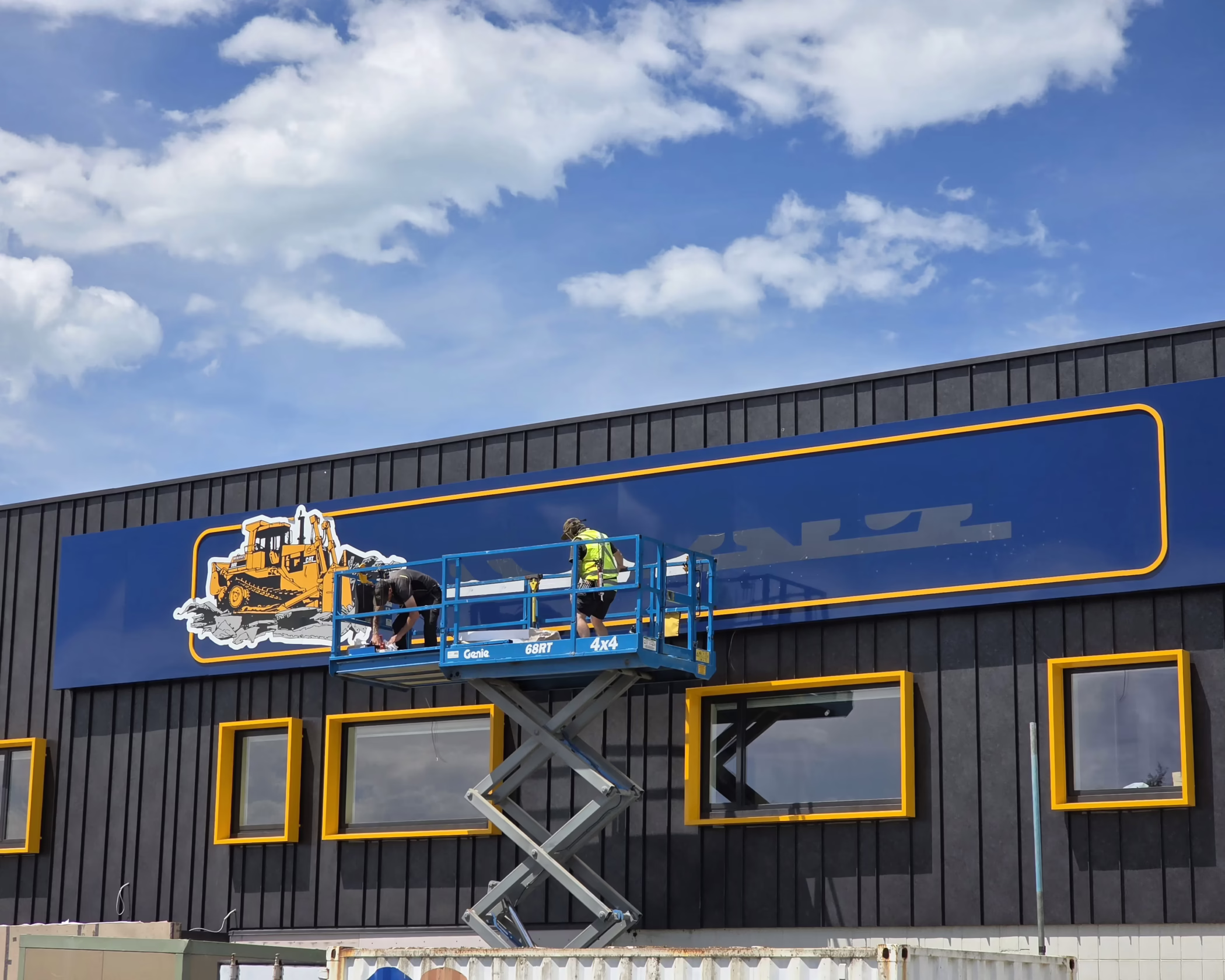 Custom exterior building fascia sign for Rooney Group, featuring a full-color graphic of an excavator. This sign is installed on a dark metal wall with bright yellow window frames, showcasing the expertise of Timaru Signs Graphix in creating custom signage solutions.