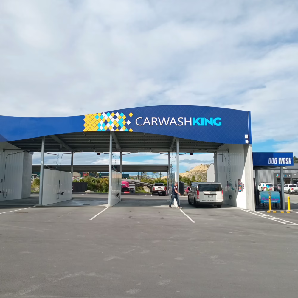 Exterior view of a Carwash King facility featuring a large blue canopy with the "CARWASHKING" logo and a geometric tile pattern, open wash bays with a parked white van, and a dedicated "DOG WASH" section to the right.