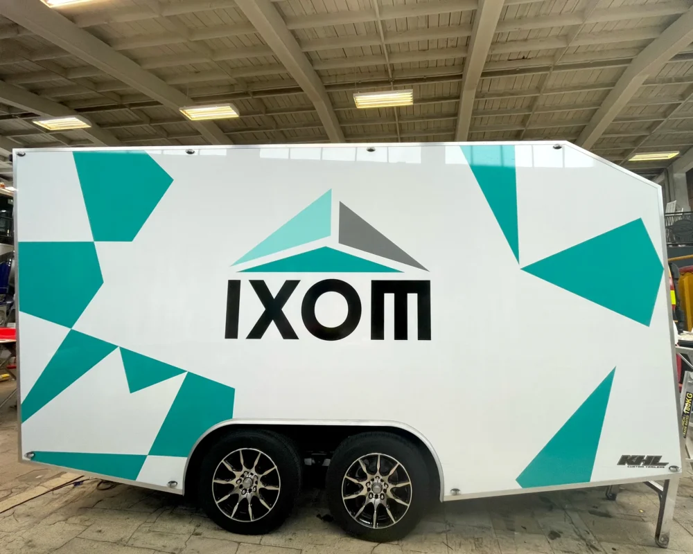 Side view of a white enclosed tandem-axle trailer featuring teal geometric graphics and the IXOM logo centered on the side panel.