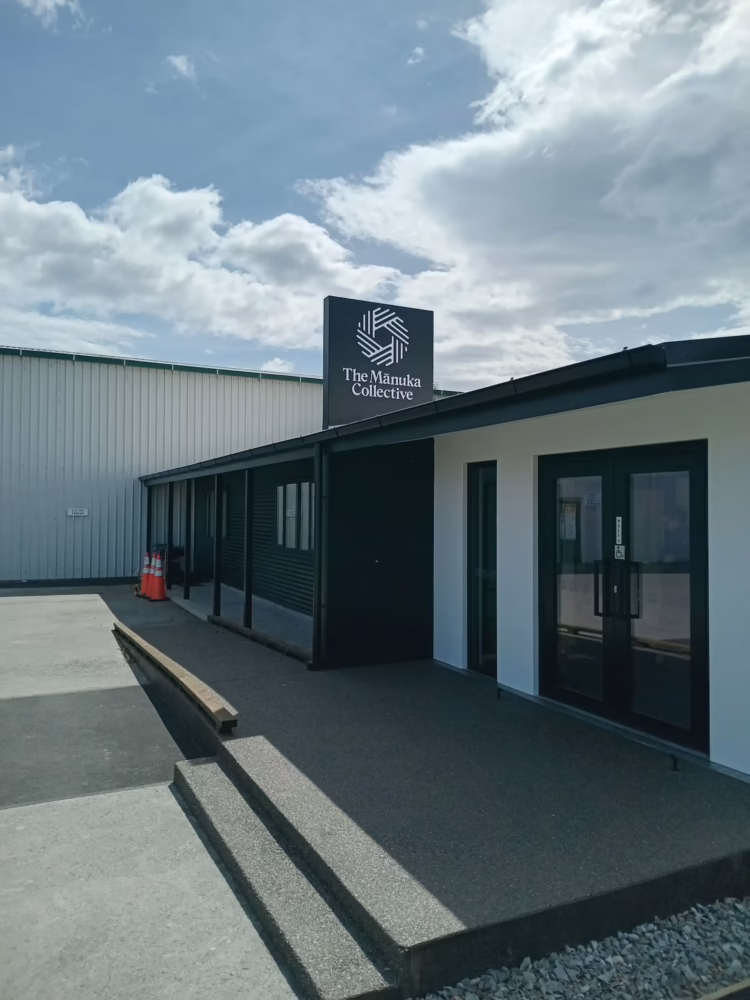 Exterior view of a modern building entrance under a cloudy blue sky, featuring dark cladding, white-trimmed glass doors, concrete steps, and a rooftop sign with a geometric logo for "The Mānuka Collective."