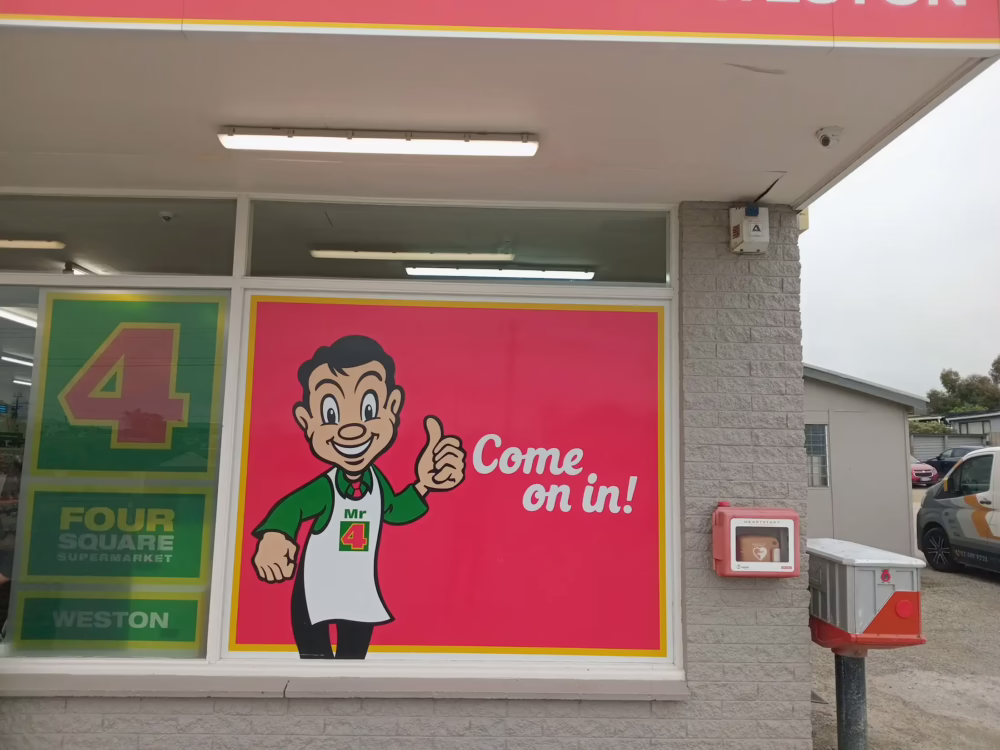 Exterior view of the Four Square Supermarket, showing a large window decal featuring the mascot, Mr. 4, giving a thumbs-up beside the text "Come on in!"