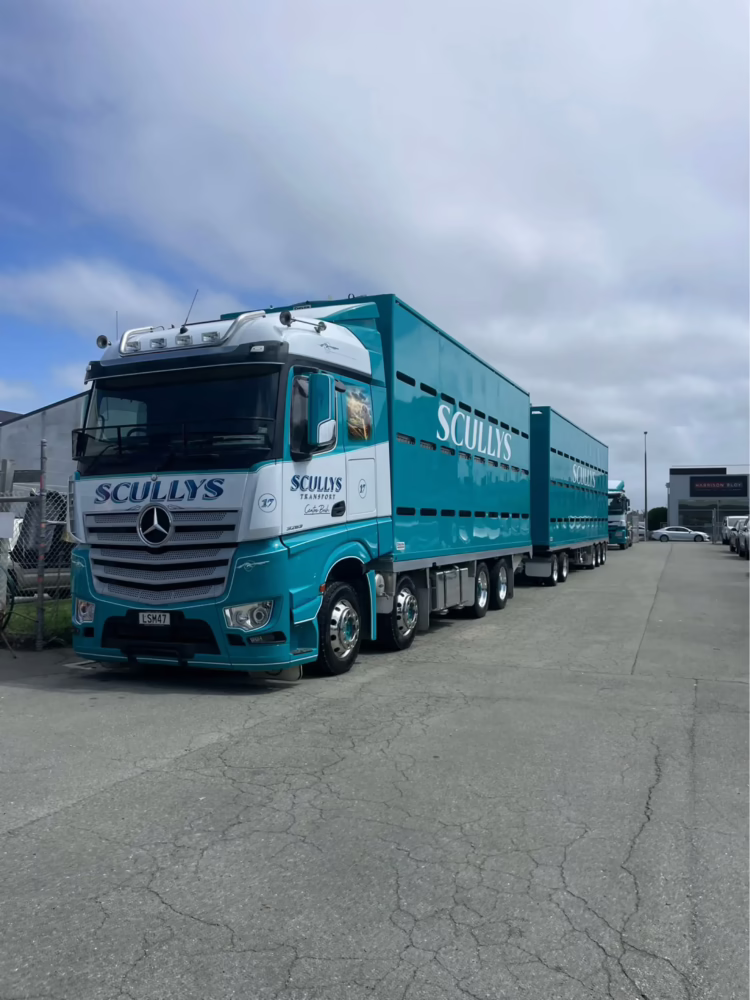 A teal and white Mercedes-Benz transport truck and trailer with "SCULLYS" branding parked in a concrete lot under a partially cloudy sky.