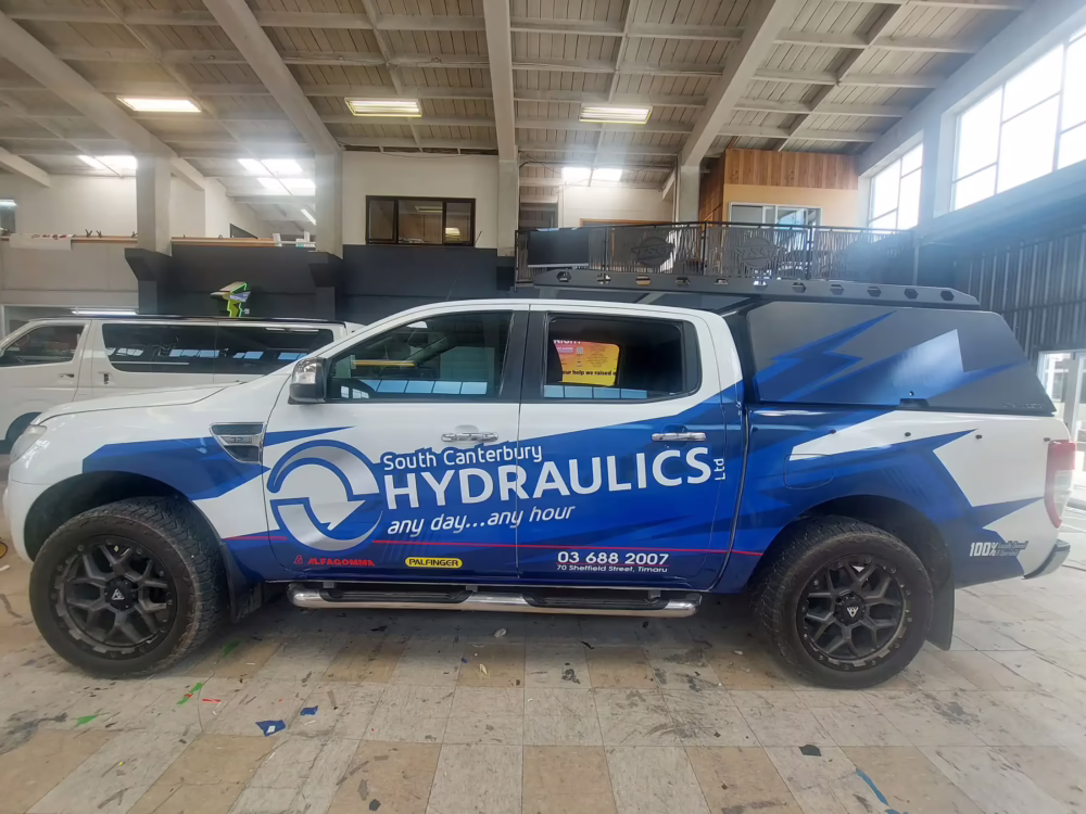 A white Ford Ranger ute with a custom blue and white South Canterbury Hydraulics vehicle wrap parked inside a signage workshop.