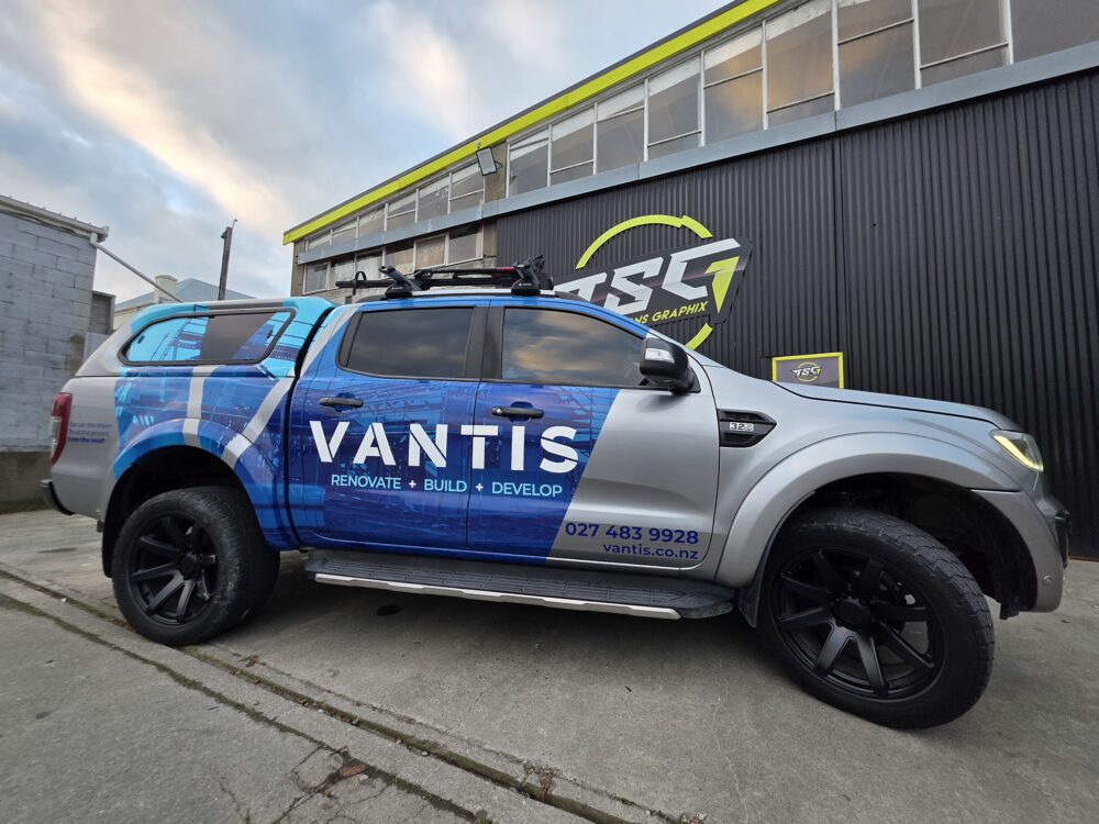 A silver double-cab Ford Ranger ute with black wheels and a rear canopy, custom-wrapped in blue graphics with white branding for 'Vantis: Renovate + Build + Develop', parked outside the Timaru Signs Graphix building.