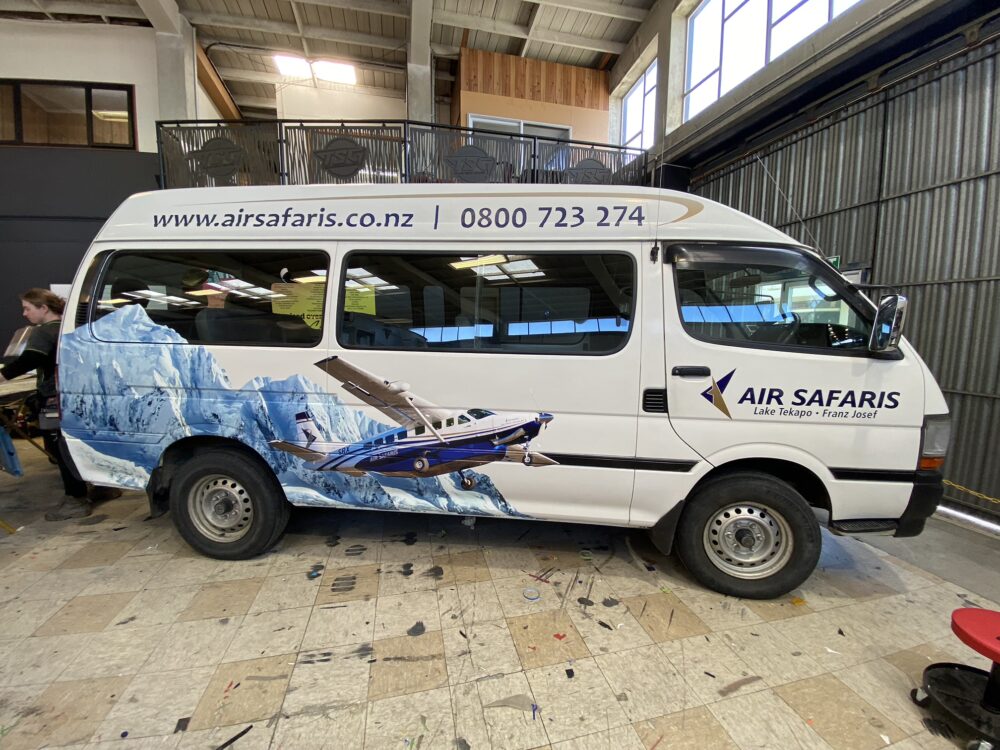 Side view of a white van parked in a workshop, featuring a vinyl wrap for Air Safaris with mountain imagery, a scenic airplane, and contact details.