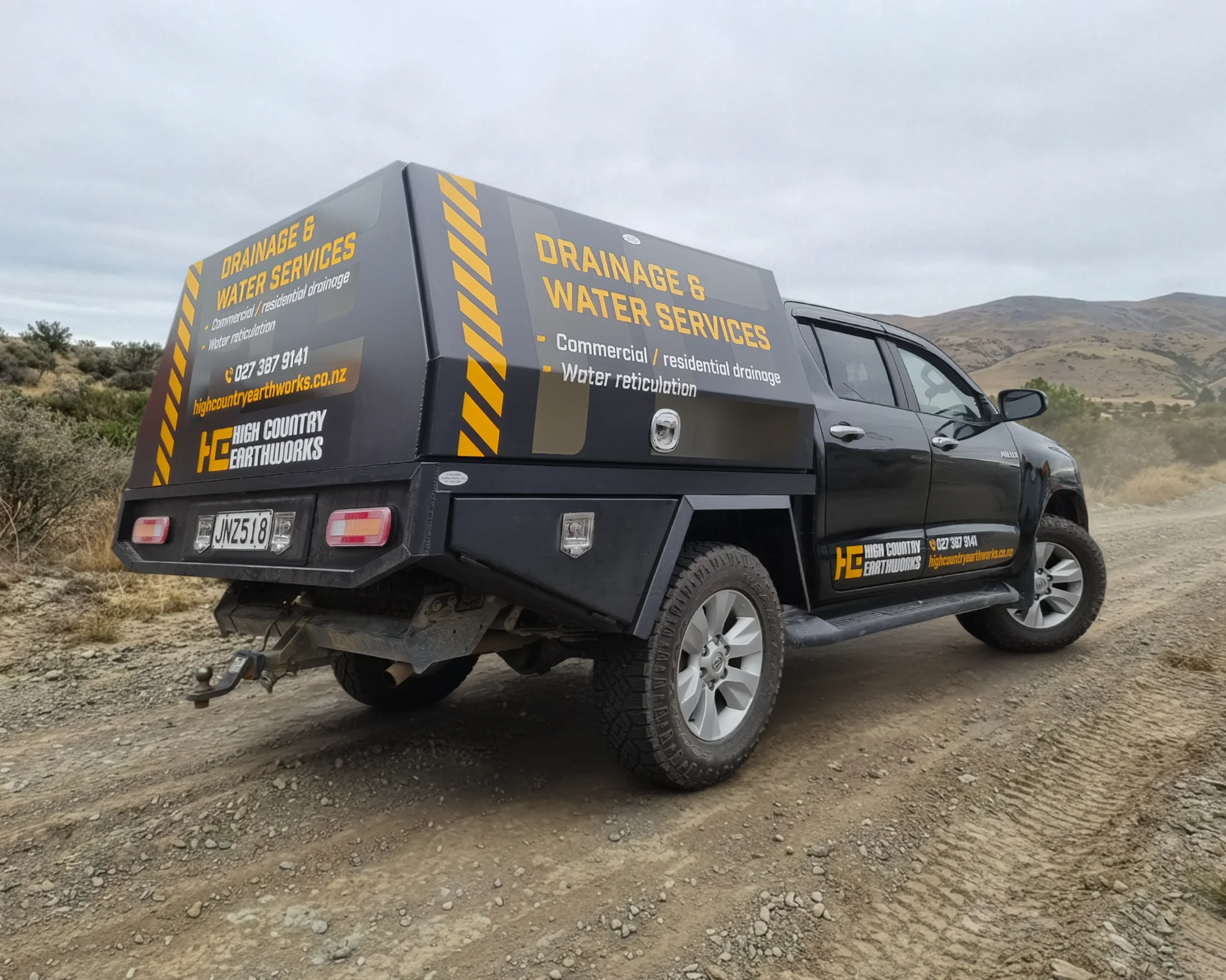 A black work ute equipped with a custom canopy featuring High Country Earthworks branding on a gravel road.