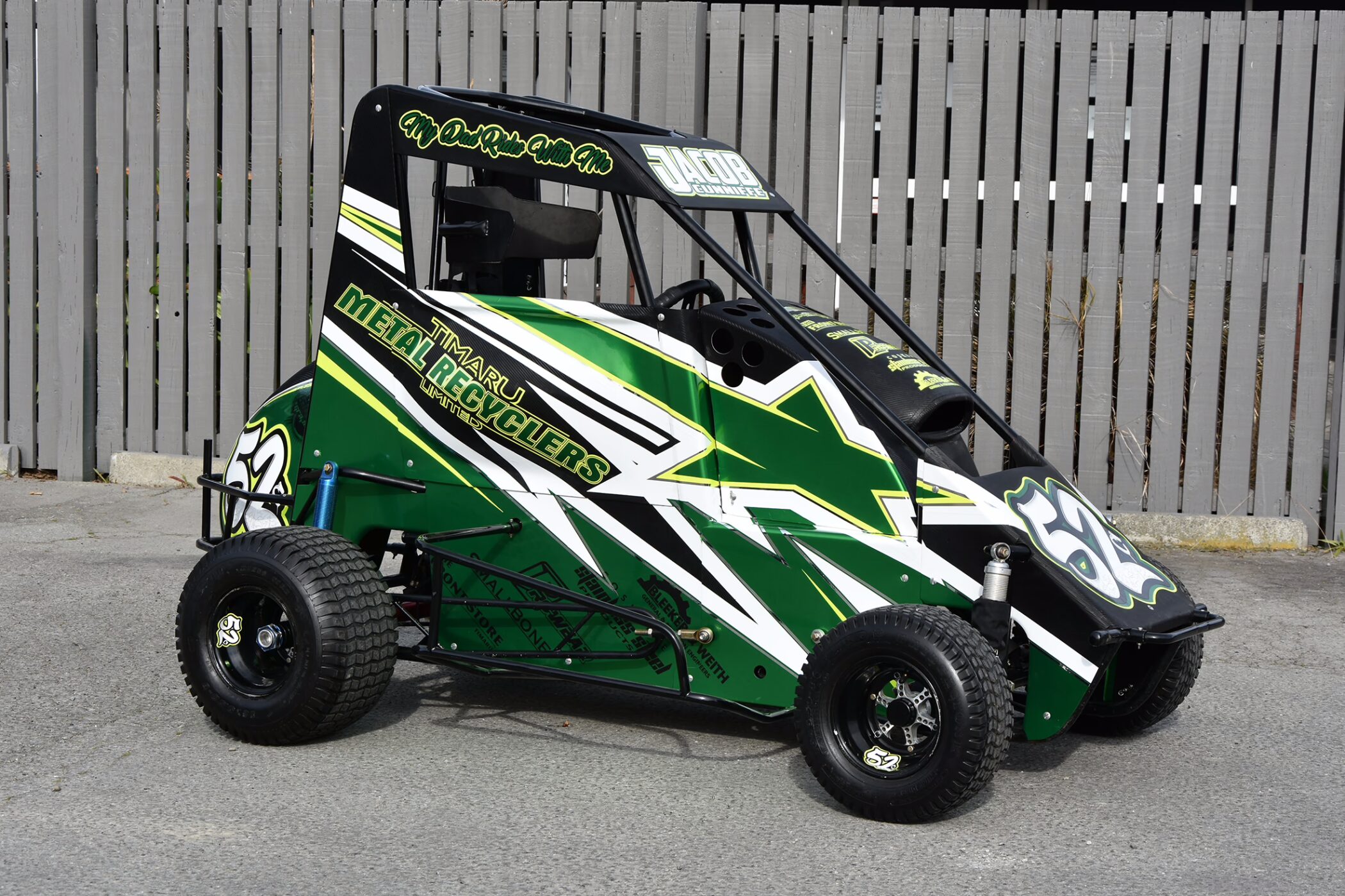 A green, black, and white quarter midget race car featuring the number 52 and custom graphics for "Timaru Metal Recyclers Limited" on its side panel. The driver's name, "JACOB CUNNLIFFE," is displayed across the top roof visor. The vehicle is parked outdoors on pavement in front of a light grey wooden fence.