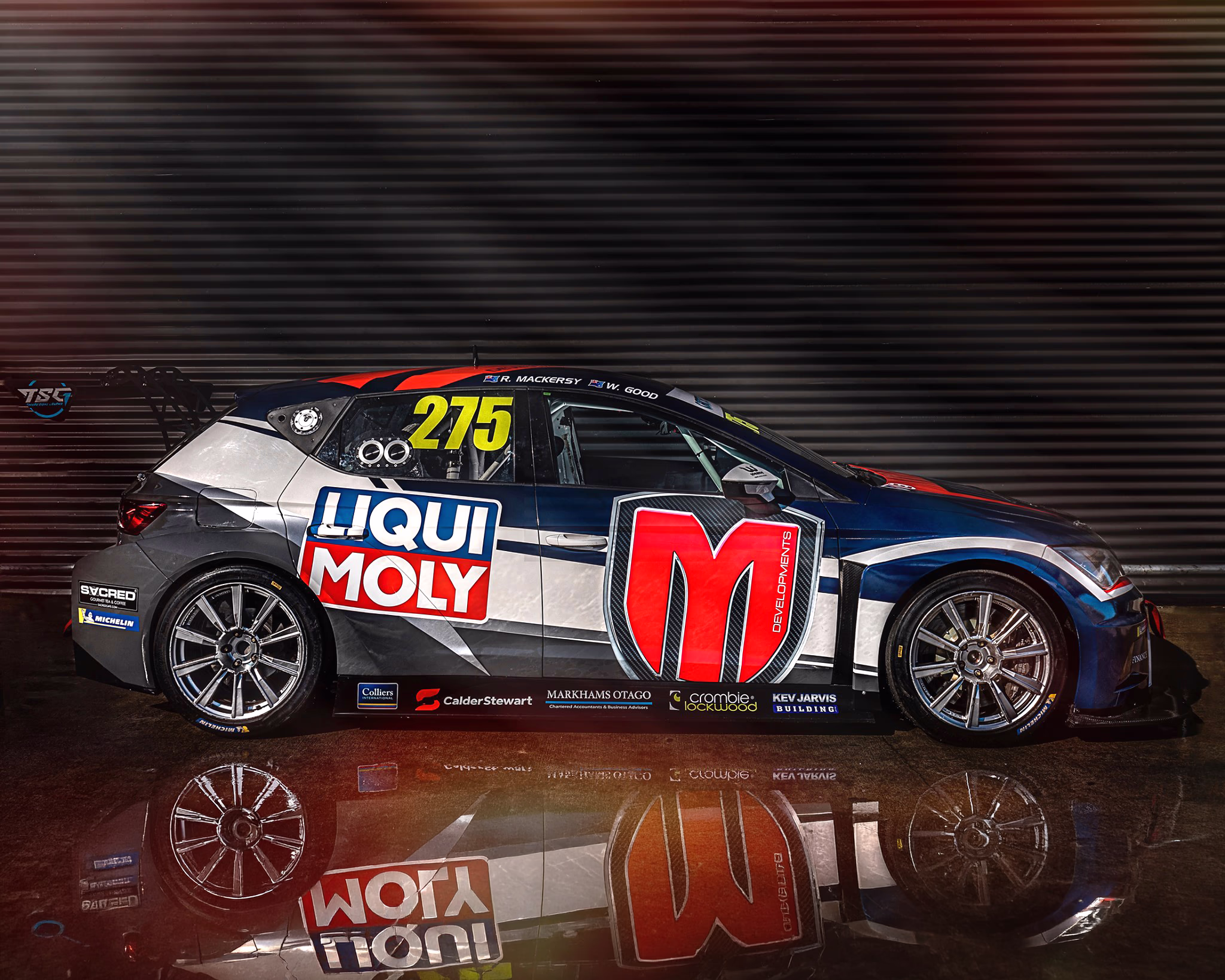 Side profile view of a blue, silver, and red race car (number 275) with custom vinyl wrap and sponsor logos—including Liqui Moly and M Developments—parked in a dark studio setting with a reflective floor.