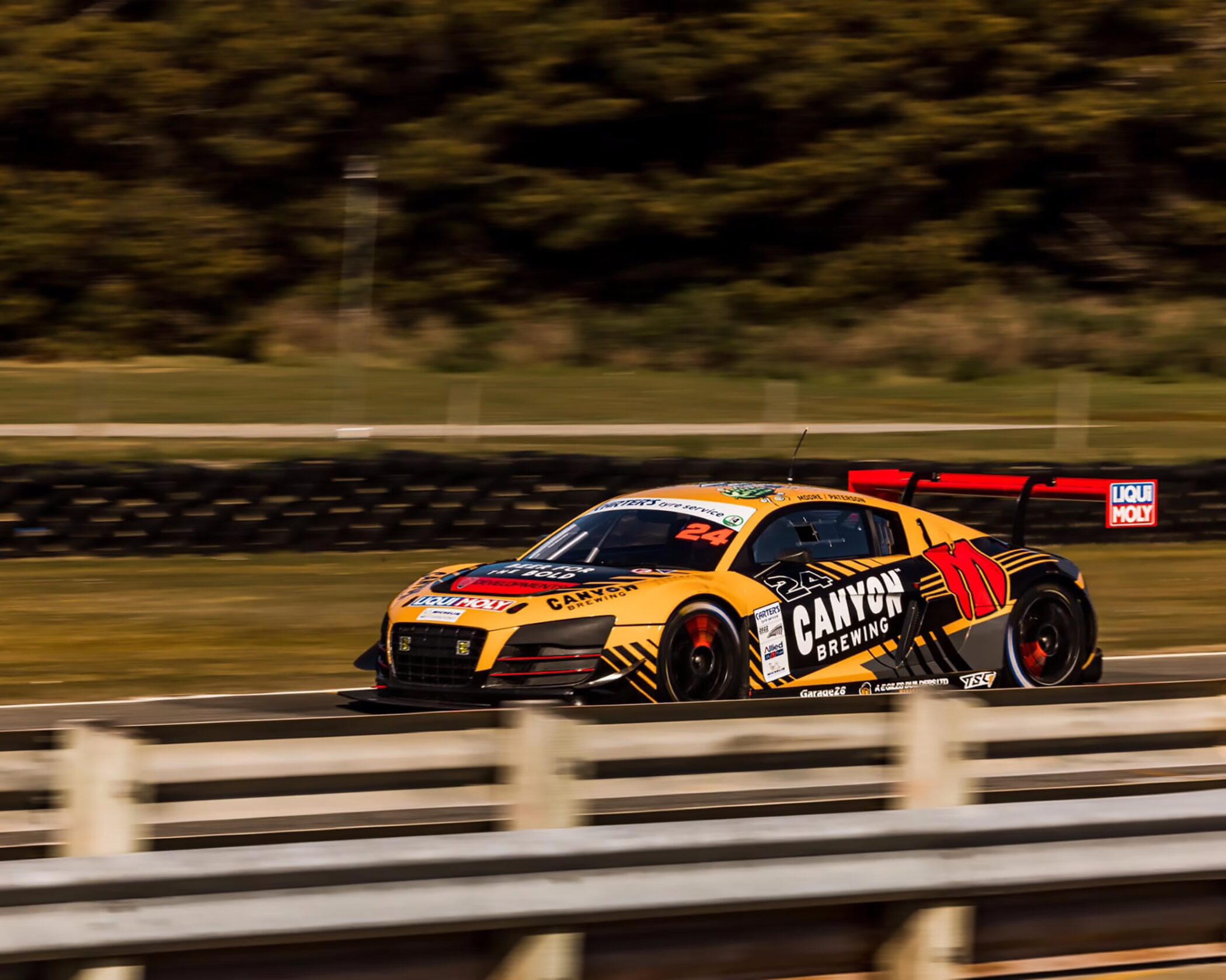 A yellow, black, and red Audi R8 LMS GT3 race car bearing number 24 and Canyon Brewing sponsorship, driving fast along a race track behind a white barrier with motion blur in the background.