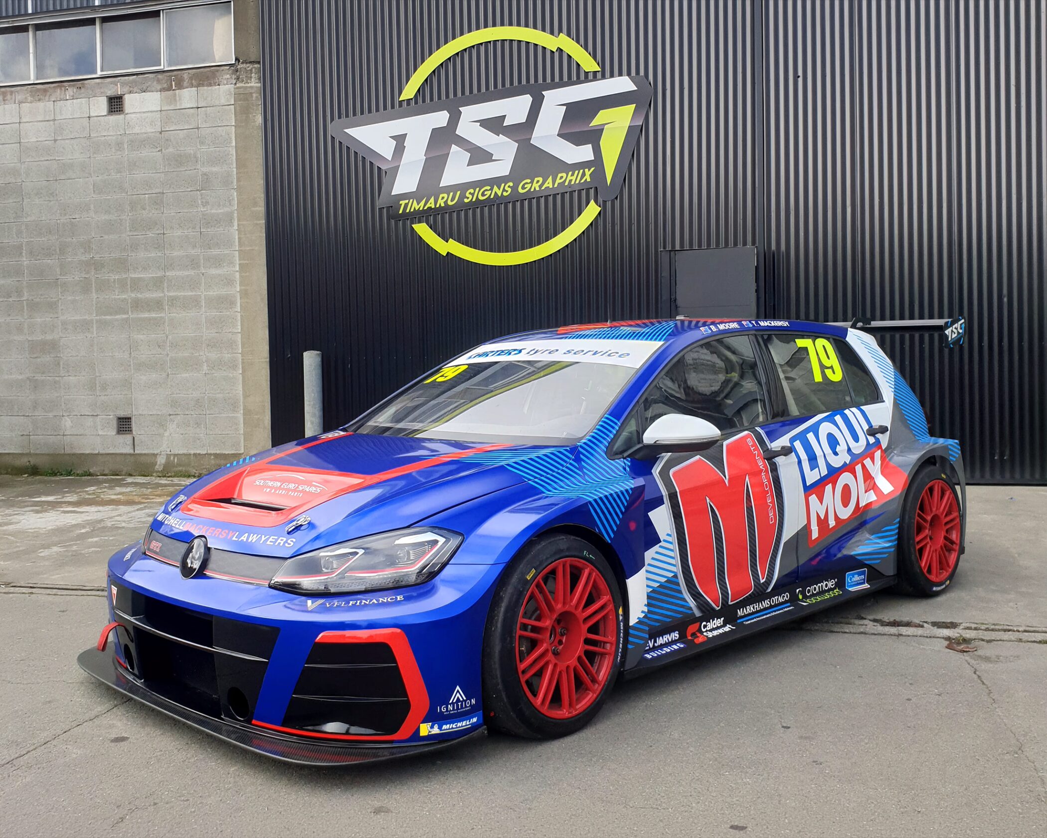 A Volkswagen Golf TCR race car featuring a vibrant blue, red, and white livery with red multispoke wheels. The car displays sponsor logos including "M Developments" and "Liqui Moly" alongside the race number 79.