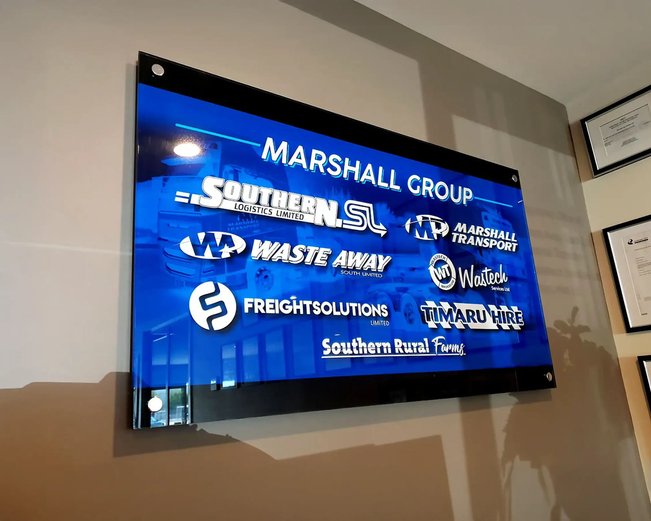 A wall-mounted glass panel displaying the "Marshall Group" logo alongside individual logos for its subsidiary companies—including Southern Logistics, Marshall Transport, Waste Away, Wastech Services, Freight Solutions, Timaru Hire, and Southern Rural Farms against a blue gradient background.