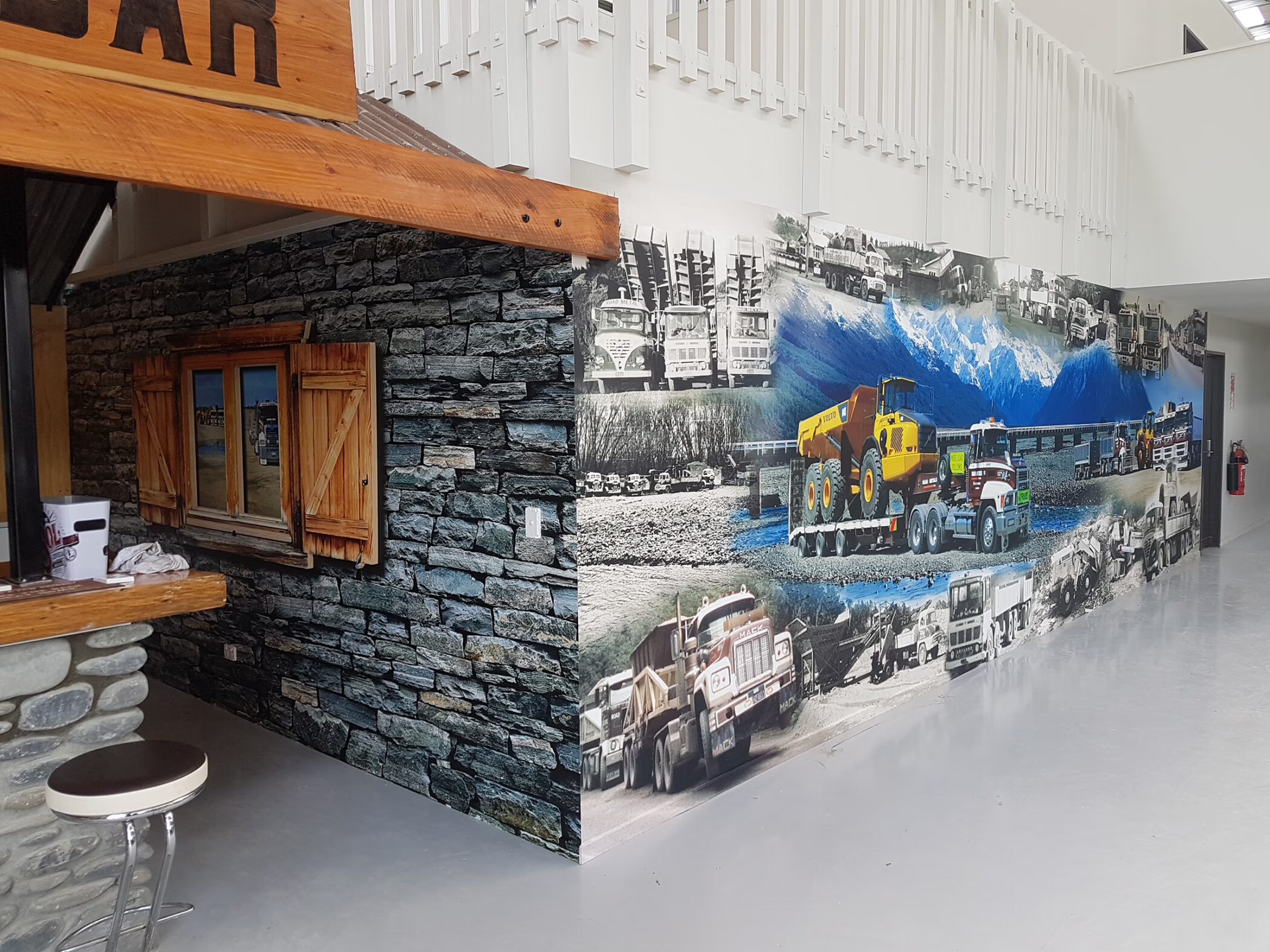 An indoor wall wrap installation featuring a rustic stone-and-timber bar facade alongside a large collage mural of heavy machinery, Mack trucks, and gravel quarry scenes.