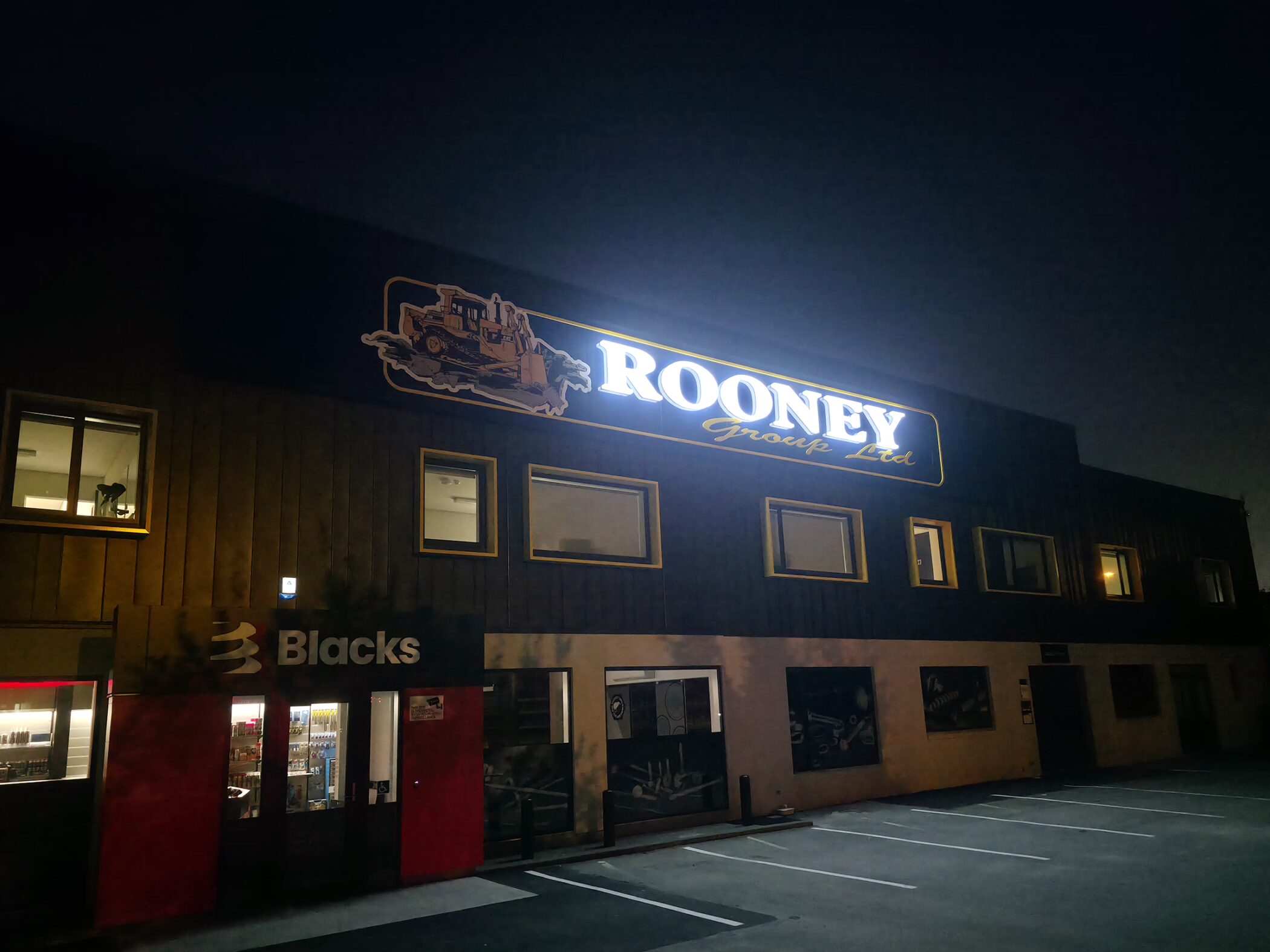 Illuminated sign for "Rooney Group Ltd" featuring a bulldozer graphic on the upper facade of a dark commercial building at night, with "Blacks" storefront visible on the ground level.