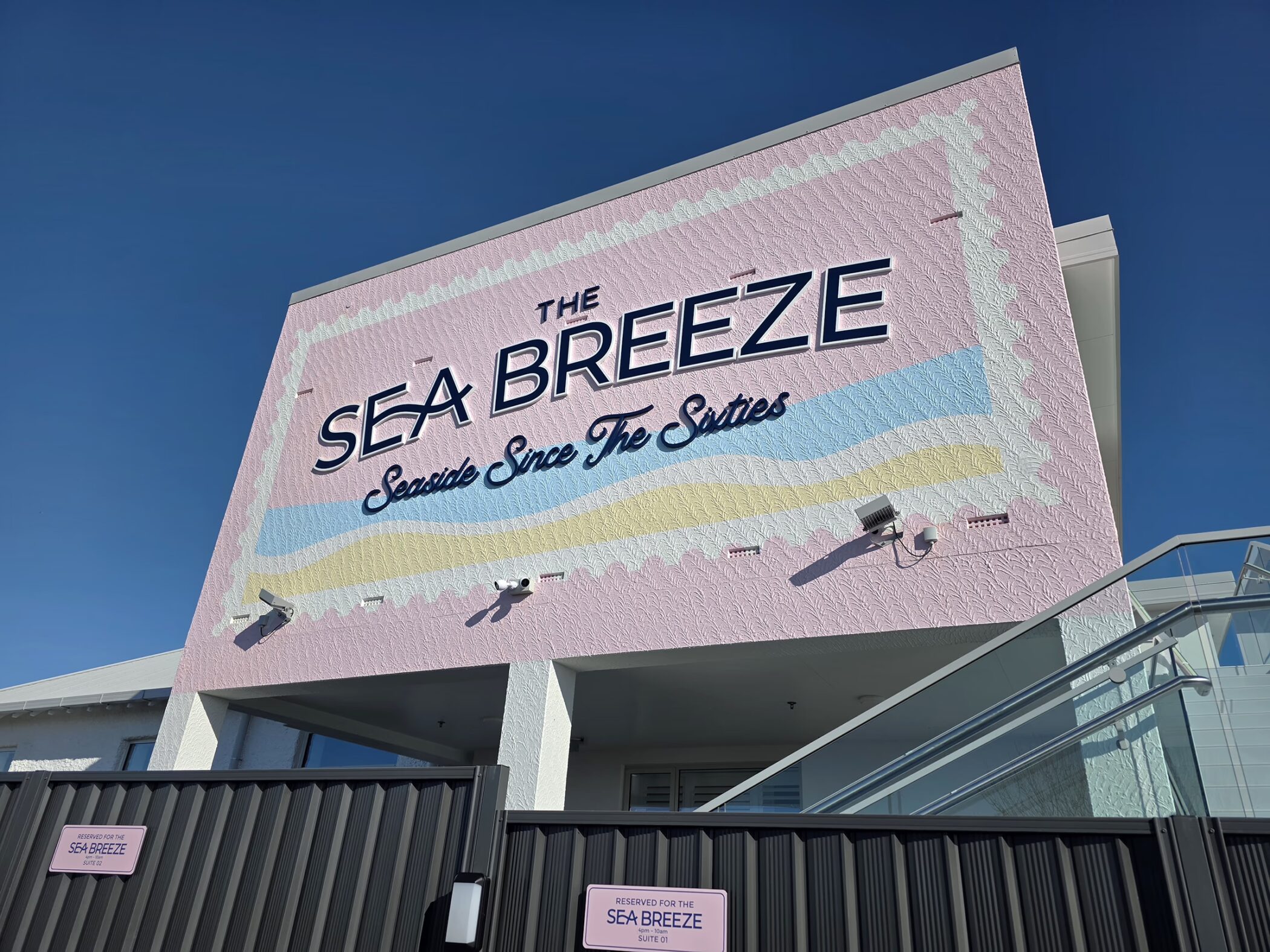 Large pink exterior building mural reading "THE SEA BREEZE - Seaside Since The Sixties" with pastel waves and stamp-style border, set against a clear blue sky.