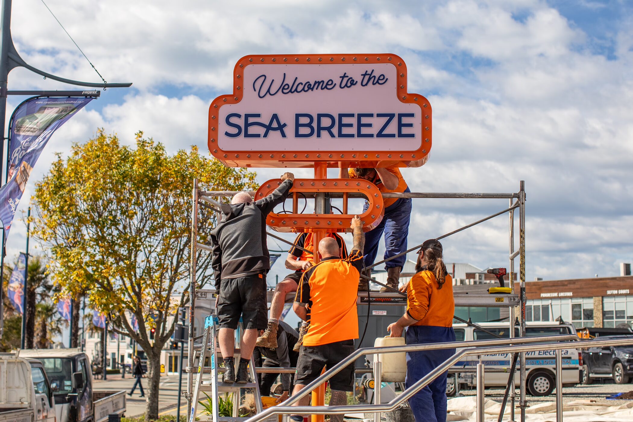 Workers on scaffolding installing a retro orange and pink sign that reads "Welcome to the SEA BREEZE".