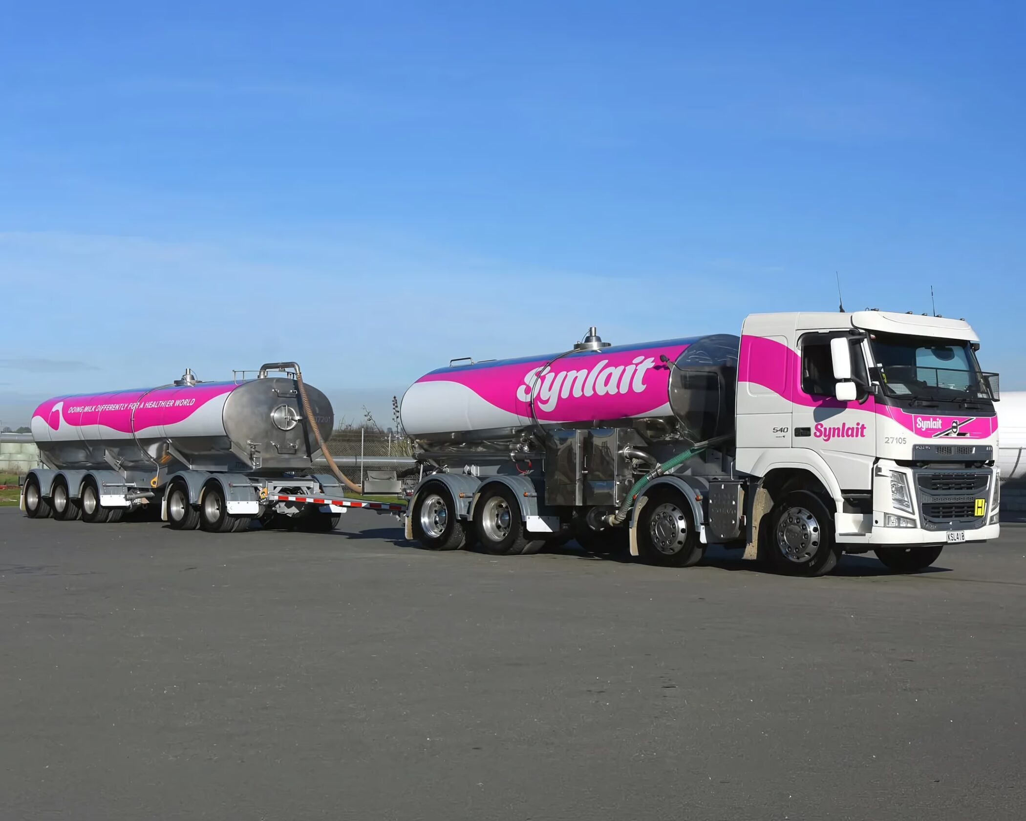 A Volvo milk tanker truck featuring bright pink Synlait branding on the tank and cab door, connected to a matching silver and pink trailer parked on tarmac.