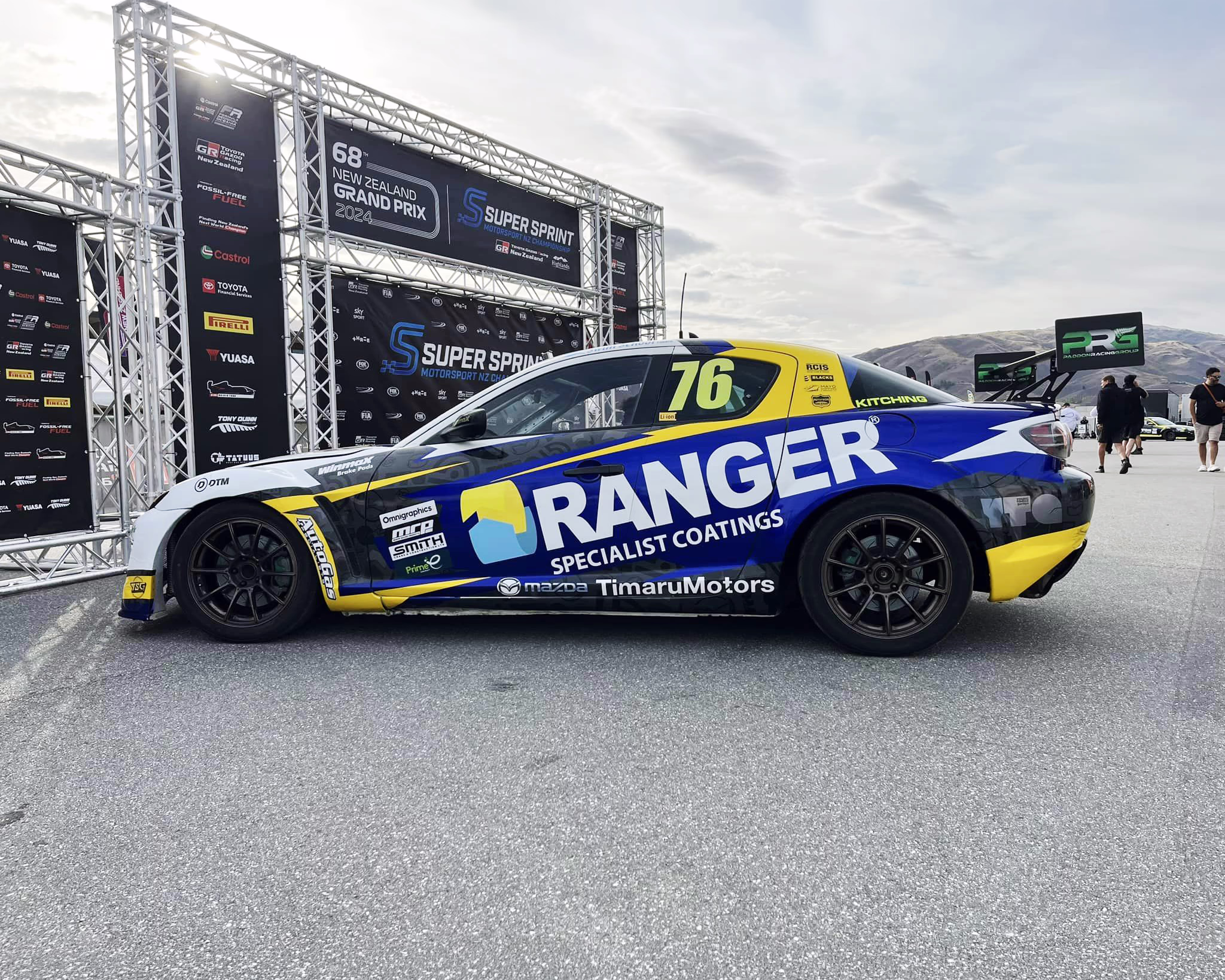 A side-profile view of a blue, yellow, and white Mazda RX-8 race car (number 76) with "RANGER Specialist Coatings" and "Timaru Motors" decals, parked on asphalt in front of a 68th New Zealand Grand Prix 2024 backdrop structure.