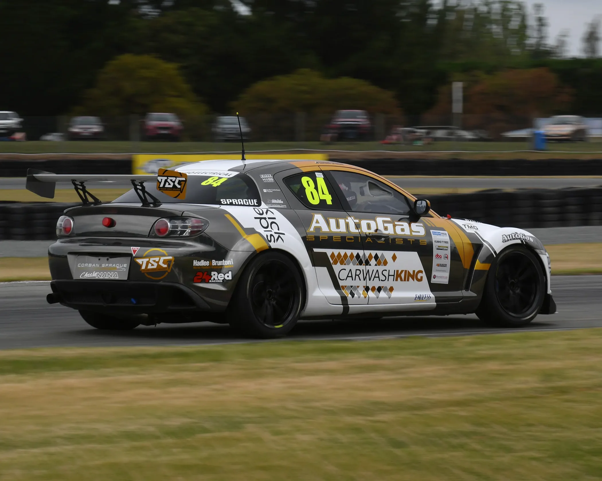 A grey, white, and gold #84 Mazda RX-8 race car driven by Corban Sprague, speeding along an asphalt circuit. The car features prominent sponsor decals including AutoGas Specialists, Carwash King, and TSC, set against a background of green grass and trees.