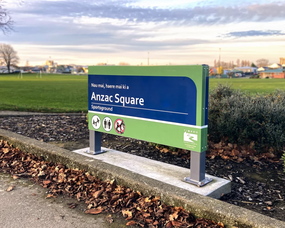 A green and blue Timaru District Council entrance sign for Anzac Square Sportsground, set beside a paved pathway next to a grassy field.