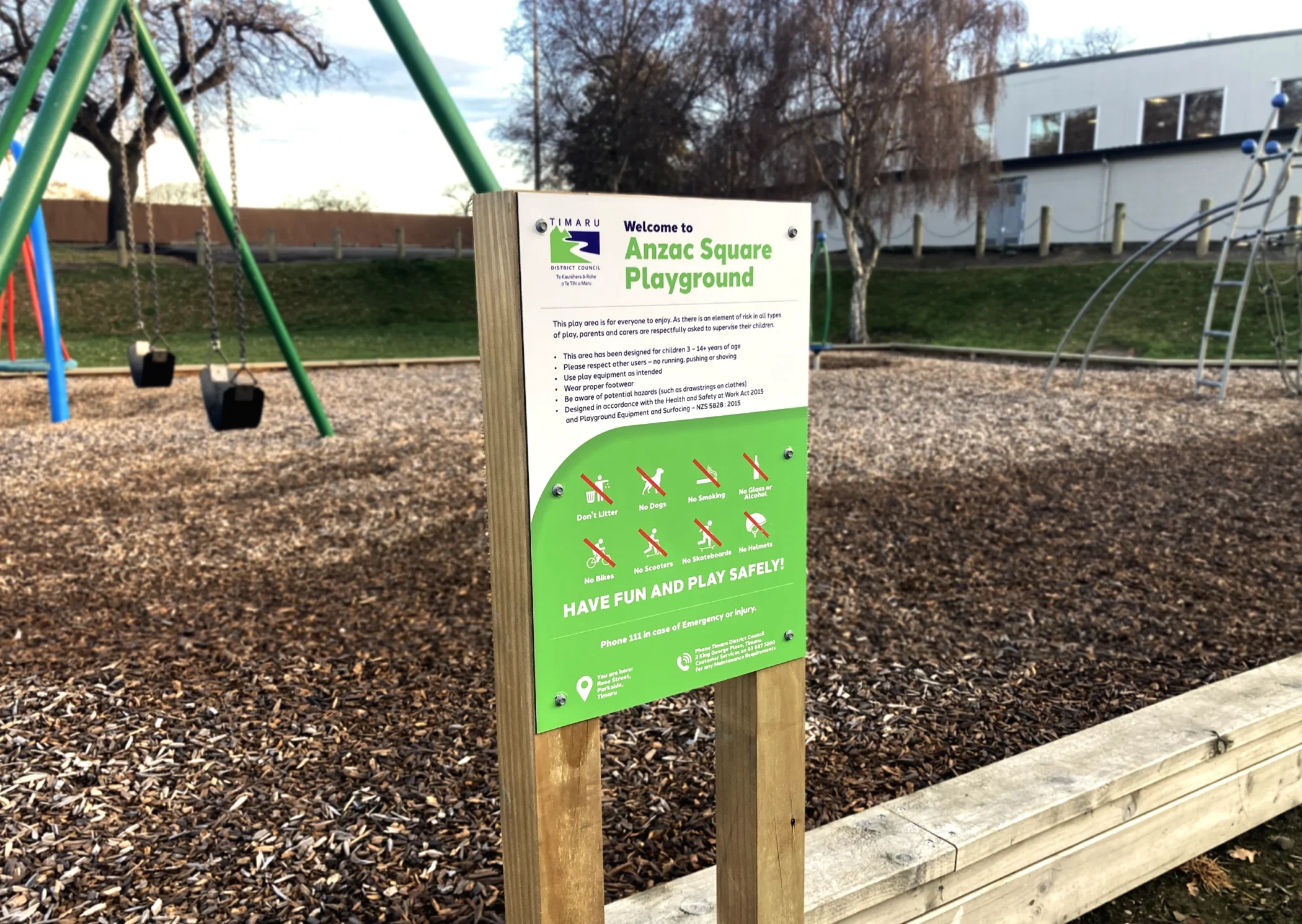 A Timaru District Council sign at the Anzac Square Playground listing rules, safety guidelines, and prohibited activities with icons, set against a background of swings and woodchips.