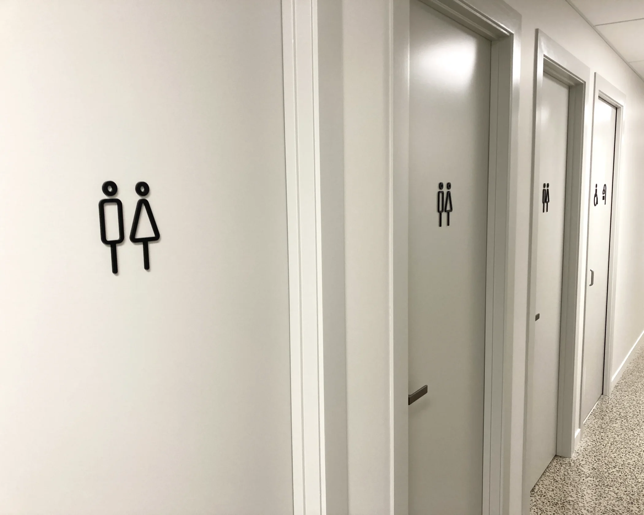 A bright white hallway featuring several toilet doors with minimal black line-art restroom icons on the doors and adjacent wall, above a light-colored speckled terrazzo floor.
