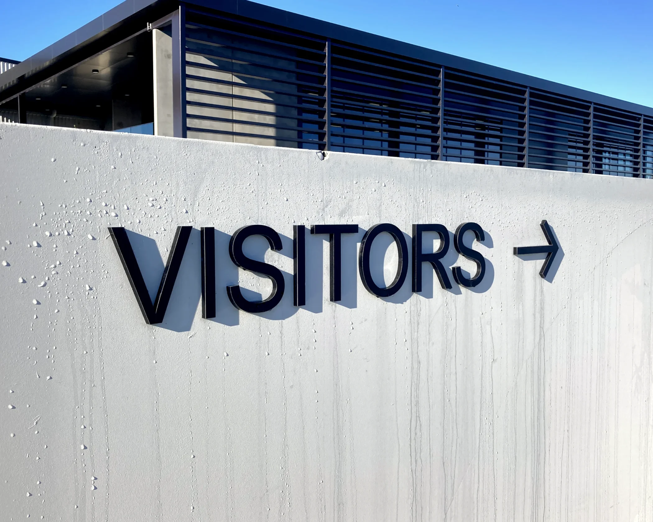 Custom black 'VISITORS →' directional sign by Timaru Signs on a concrete building with blue louvers.