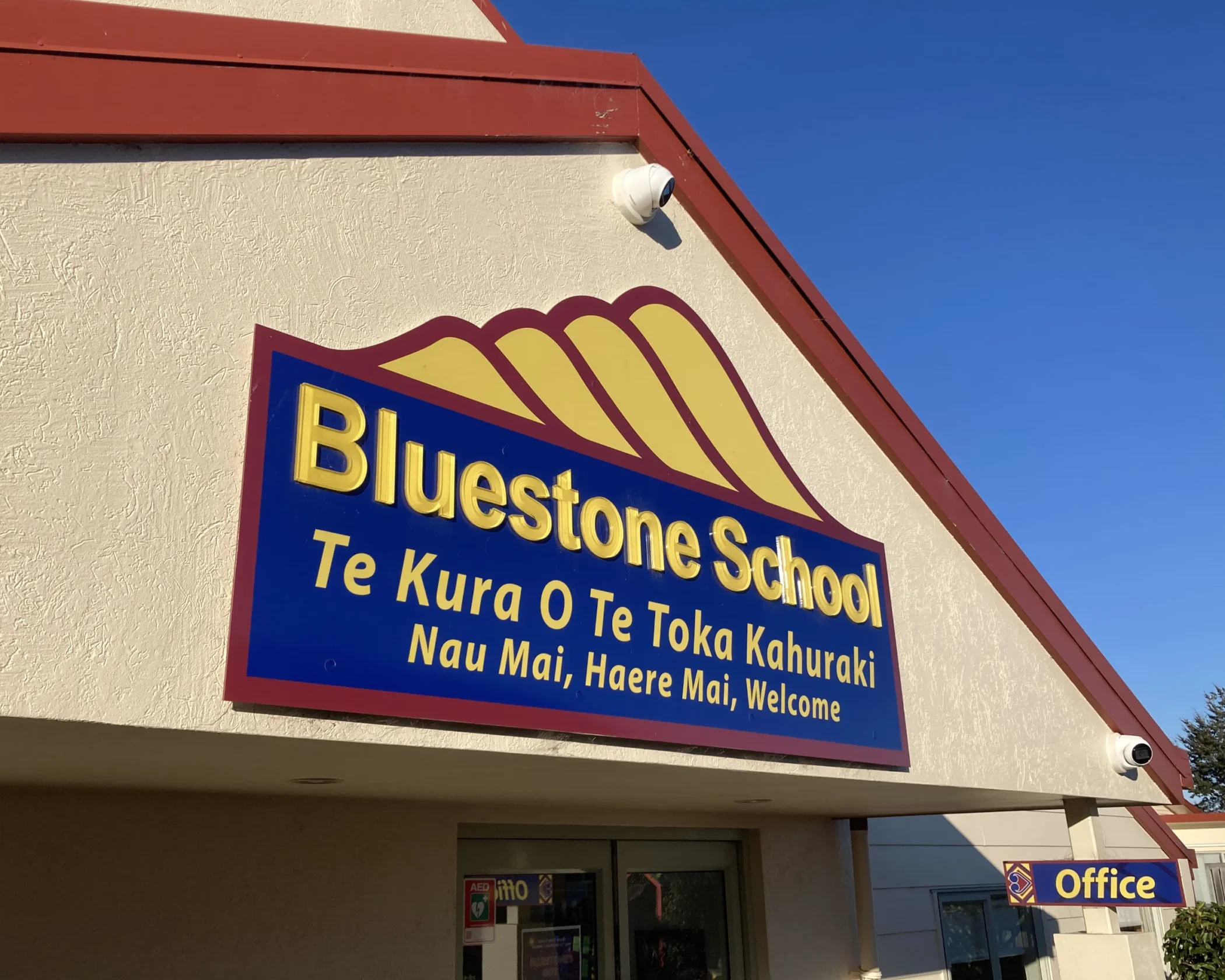 Front entrance sign of Bluestone School reading "Bluestone School - Te Kura O Te Toka Kahuraki - Nau Mai, Haere Mai, Welcome" with an "Office" sign below.