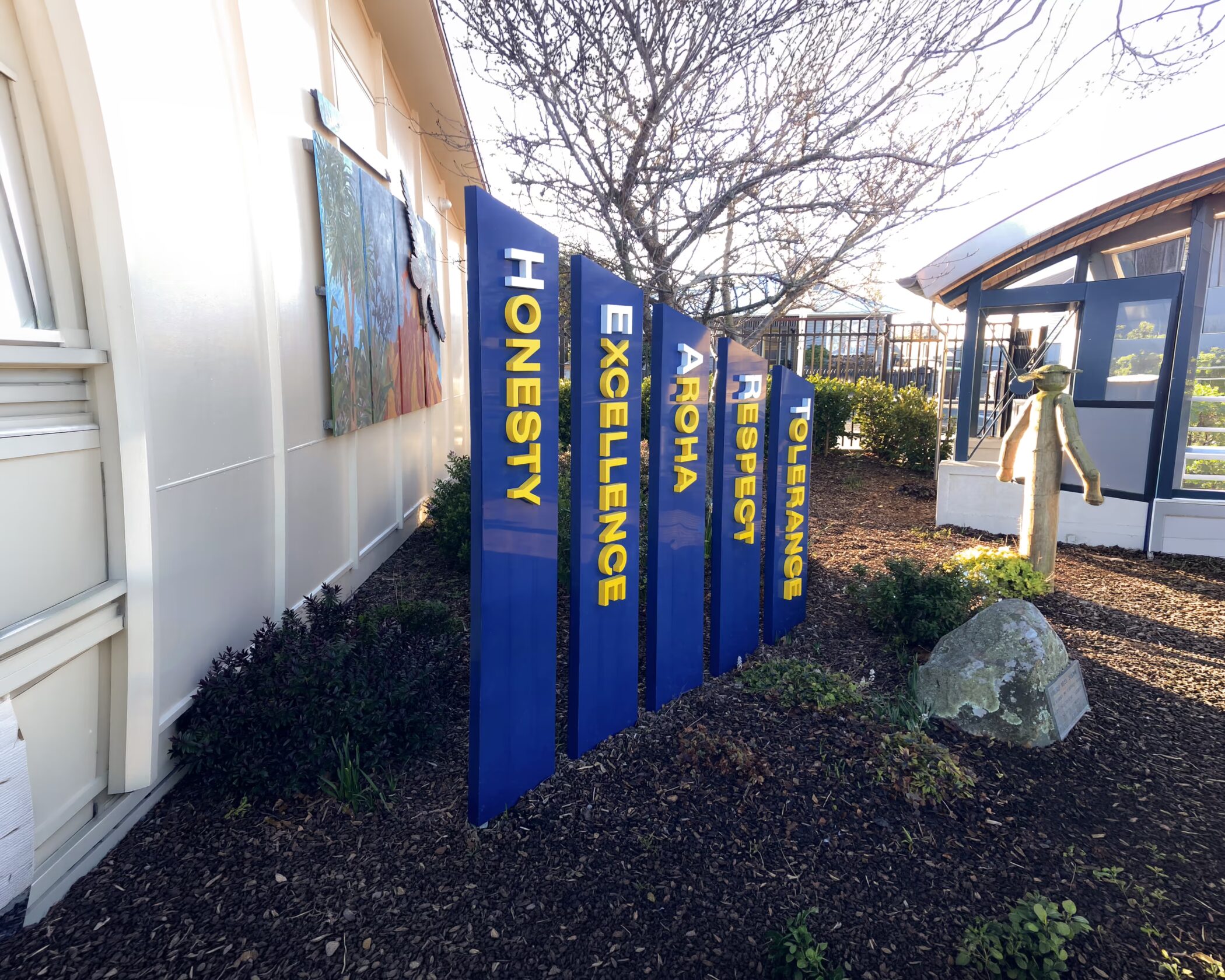 Blue signposts displaying school / organizational values—HONESTY, EXCELLENCE, AROHA, RESPECT, and TOLERANCE—printed in yellow text, set in a landscaped garden area next to a building.