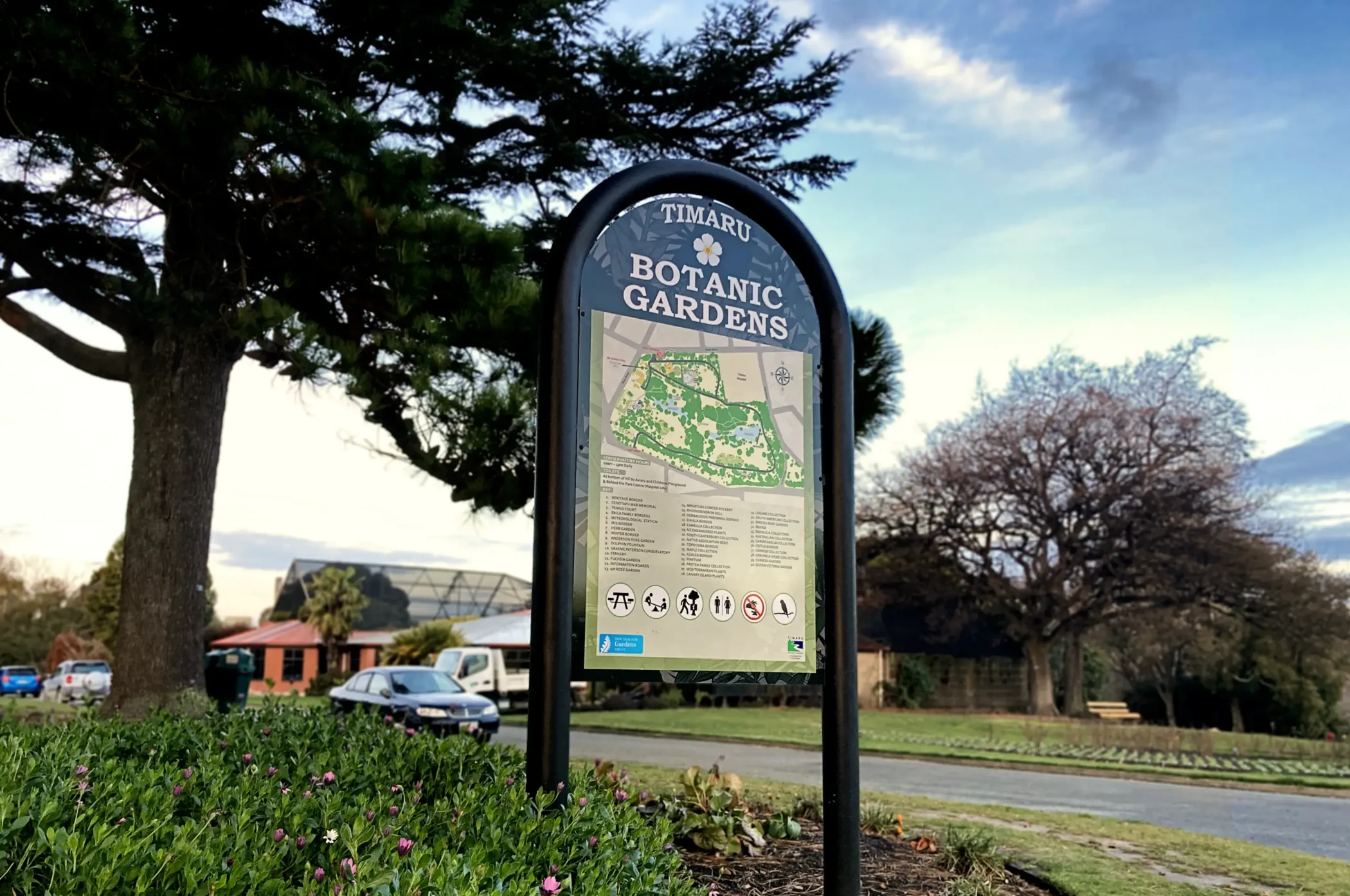 An outdoor sign for the Timaru Botanic Gardens displaying a map of the grounds and a numbered legend, set against a background of grassy lawns, trees, and parked cars under a clear sky.