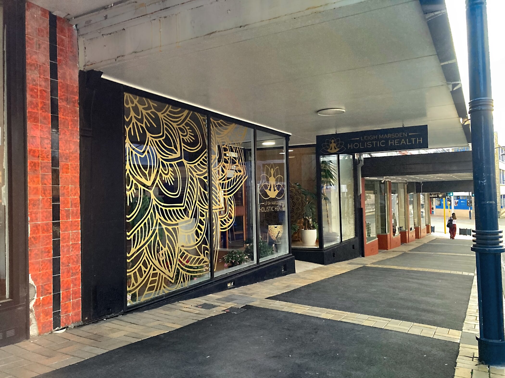 Exterior view of Leigh Marsden Holistic Health shopfront featuring gold decorative line-art window decals and a branded overhead sign under a covered arcade walkway.