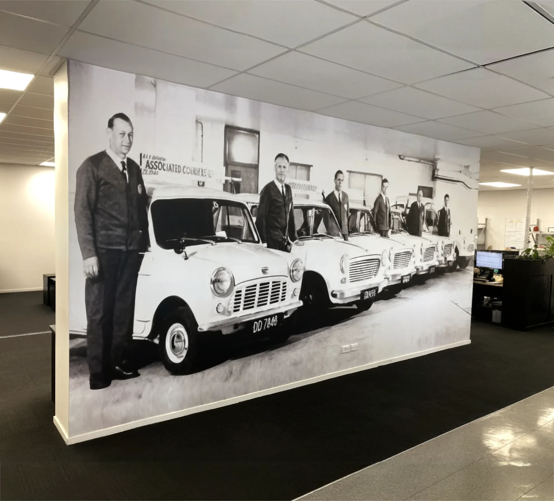 Black and white historical photo printed as a full-wall mural inside a modern office, featuring line-up of drivers standing next to vintage courier cars.