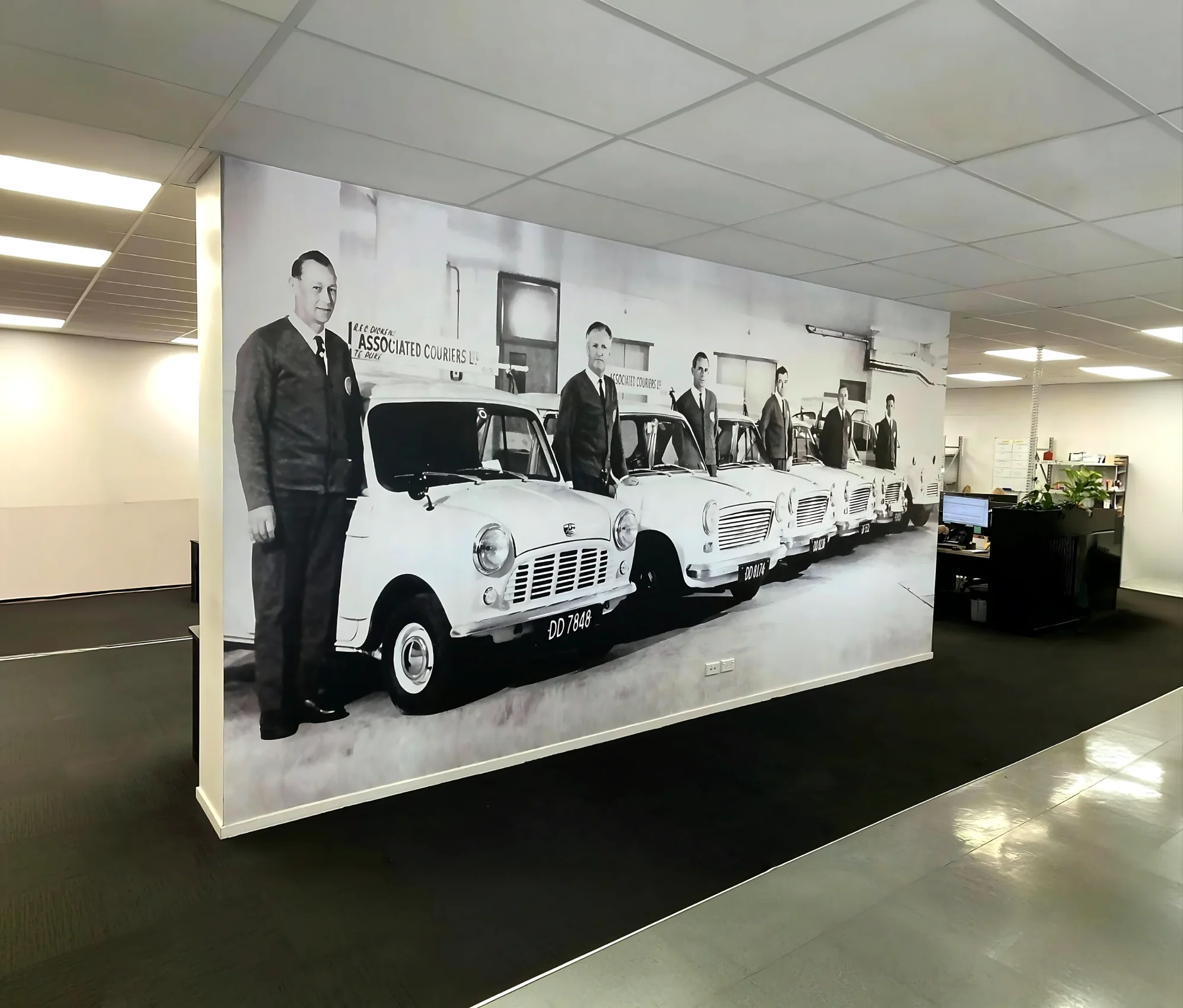 Black and white historical photo printed as a full-wall mural inside a modern office, featuring line-up of drivers standing next to vintage courier cars.