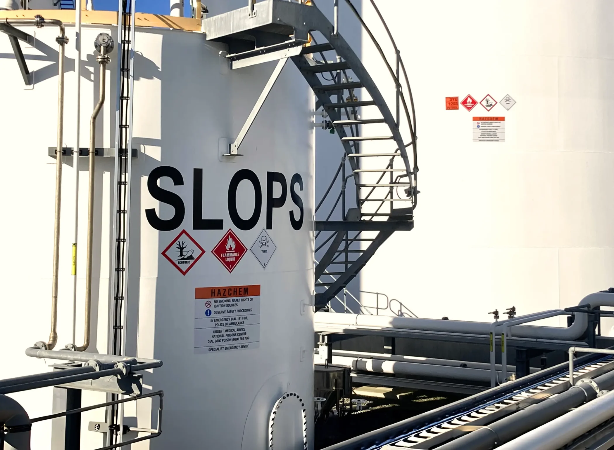 Large "SLOPS" lettering, HAZCHEM warnings, and GHS icons on a white industrial fuel tank with complex piping at Tasman Fuels.